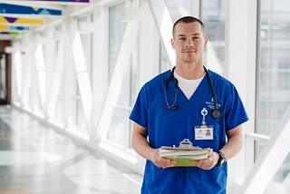 A BSN nurse in scrubs, holding a clipboard and wearing a stethoscope in the corridor of a hospital.