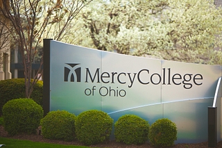 Metal sign that says Mercy College of Ohio surrounded by landscaping including bushes and flowering trees.